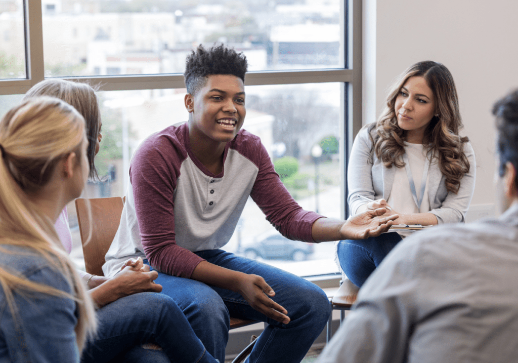 An African teenager speaking and sharing personal stories during a Mental, Emotional, and Social Health (MESH) group session on a college campus, promoting emotional well-being and peer support.