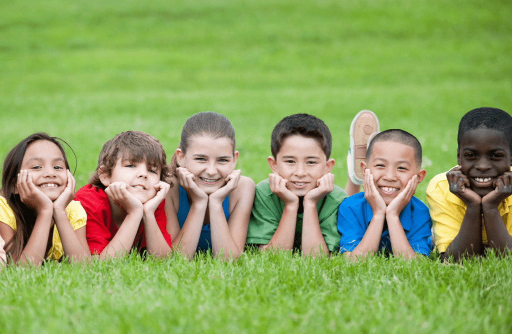 Diverse-Kids-Playground-Sports-Camp A diverse group of boys and girls with various skin tones smiling and looking at the camera while on a playground at a sports camp, capturing a moment of joy and togetherness.