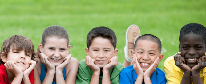 A diverse group of boys and girls with various skin tones smiling and looking at the camera while on a playground at a sports camp, capturing a moment of joy and togetherness.