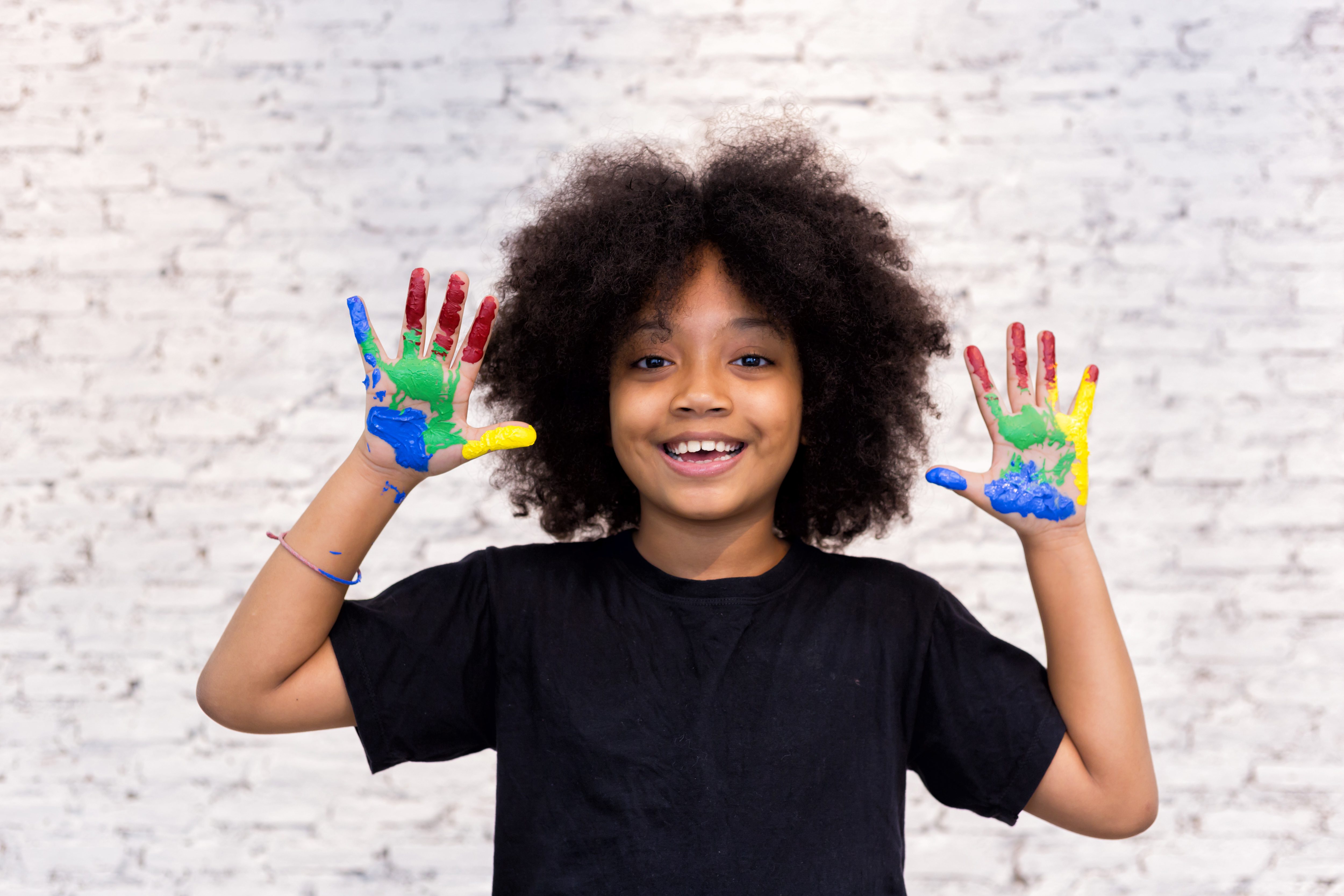 African American playful and creative kid getting hands dirty with many colors – in white brick background.